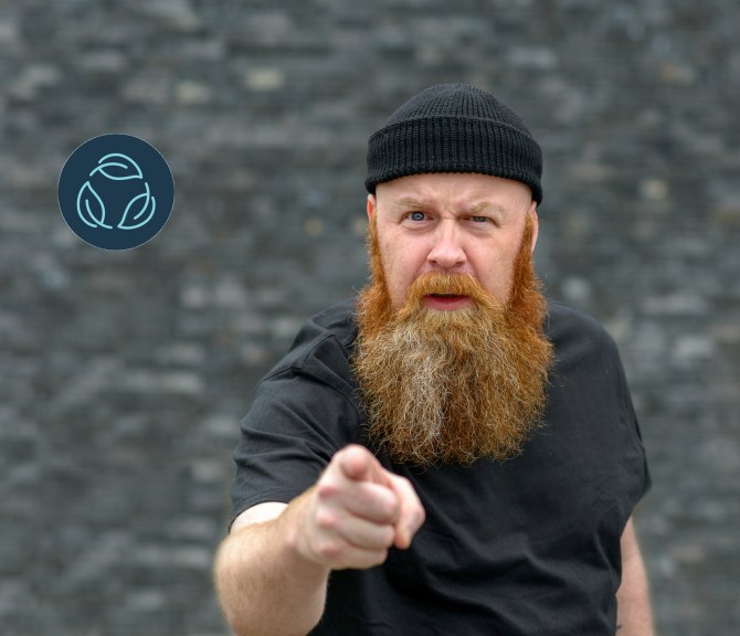 A bearded person points at the camera, gray background.