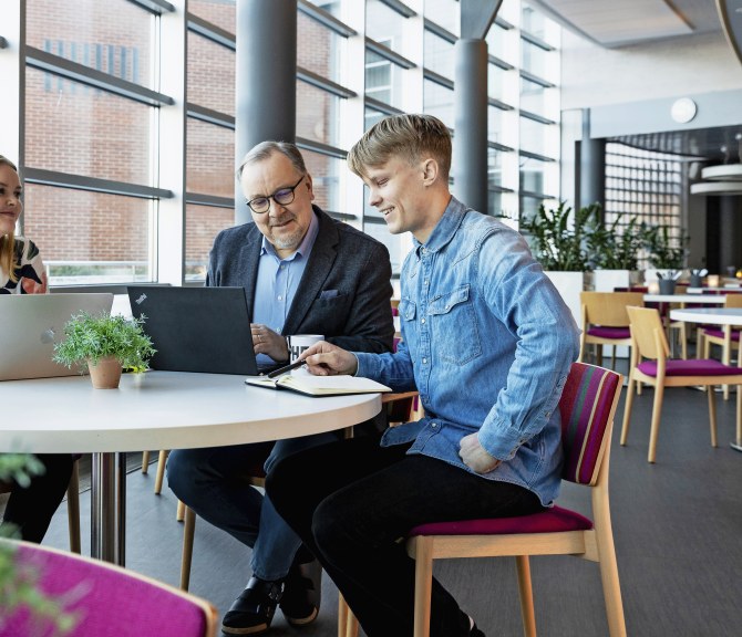 Kolme ihmistä istuu saman pöydän ääressä ruokalassa, edessään kaksi kannettavaa tietokonetta ja vihko.