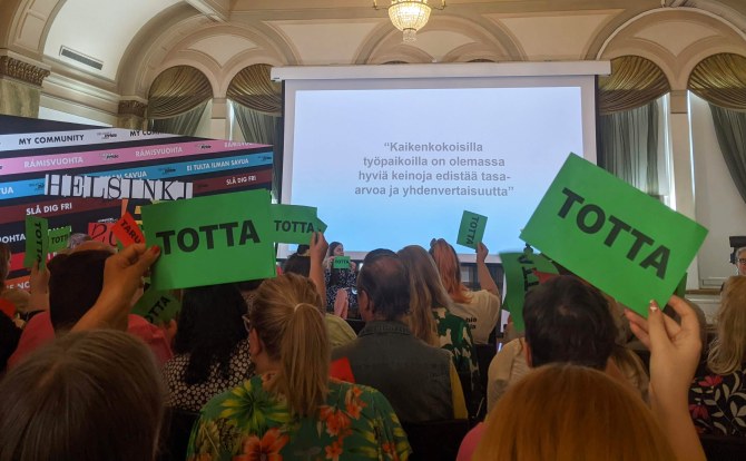 Pride-seminaarin yleisö on nostanut ilmaan vihreitä totta-lappuja, taustalla näkyy teksti: kaikenkokoisilla työpaikoilla on olemassa hyviä keinoja edistää tasa-arvoa ja yhdenvertaisuutta.