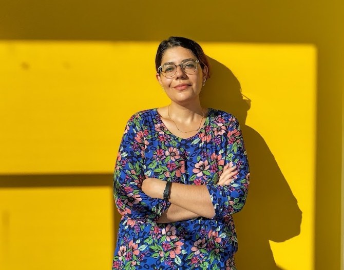 Negar Sharifi leaning on a sun-lit yeallow wall.