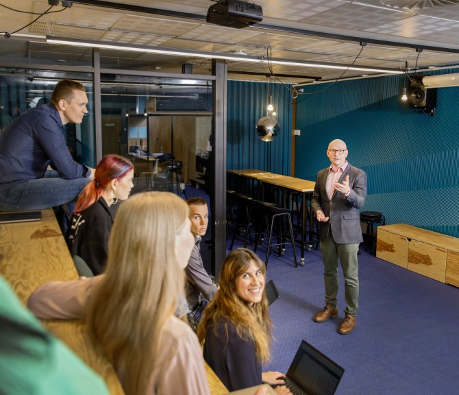 Joukko henkilöitä istuu TEK Loungen katsomossa, yksi puhuu edessä.