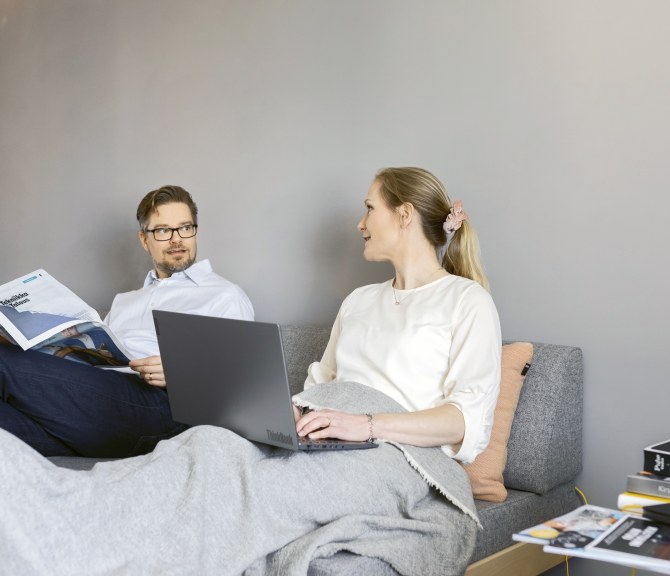 Kaksi henkilöä istuu sohvalla, toinen lukee lehteä ja toisella on tietokone edessään.