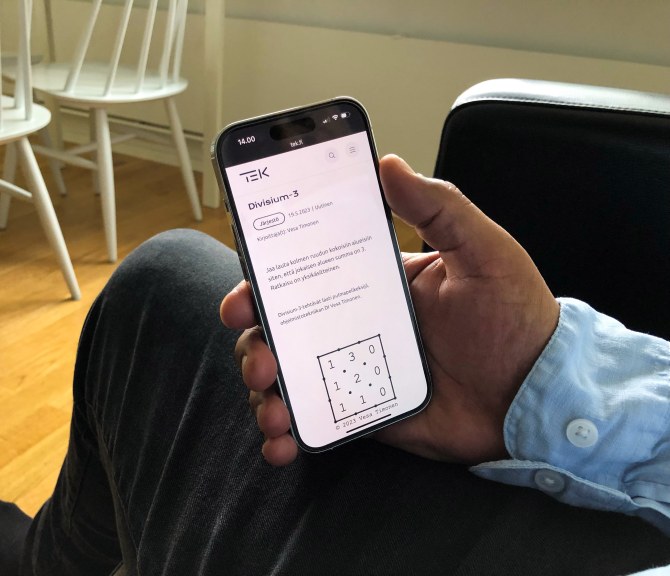 Kotioloissa istuva henkilö pitää kädessään kännykkää, jossa on auki Divisium-3-peli.