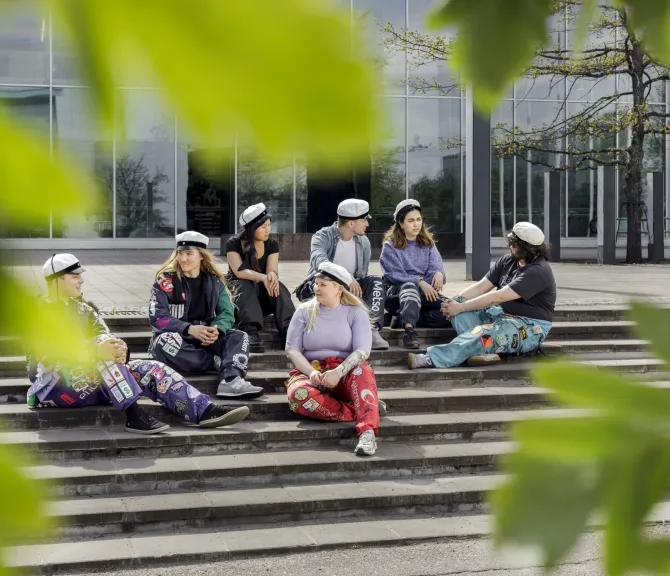 Opiskelijoita istumassa ulkona portaikossa, kesän vihreitä lehtiä etualalla.