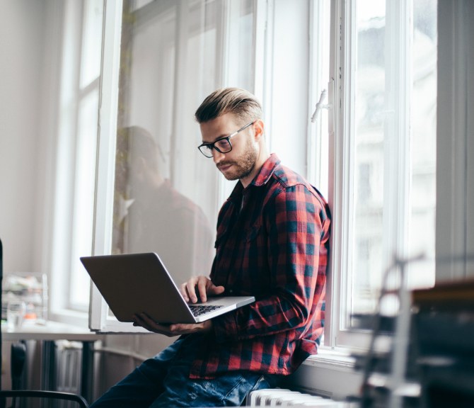 Ruutupaitainen mies istuu läppärin kanssa toimiston ikkunalaudalla.
