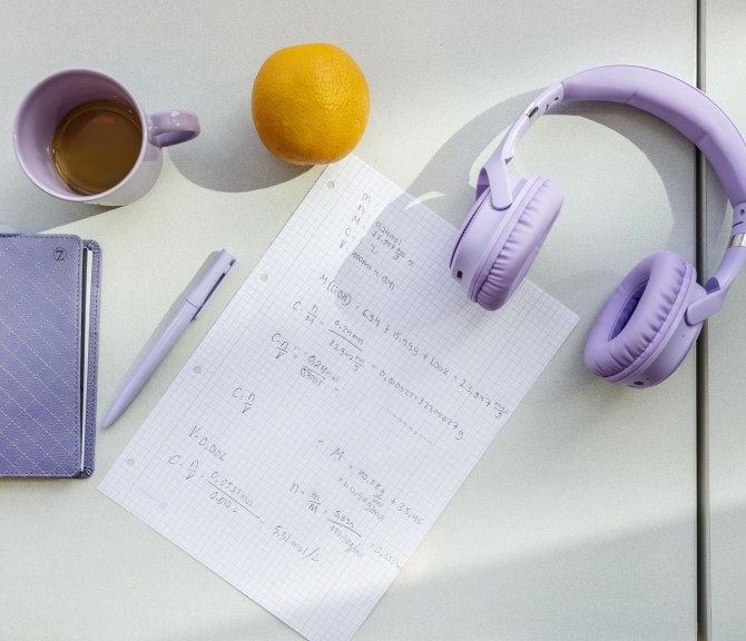 Pöydällä kuulokkeet, kahvikuppi, kalenteri, kynä, appelsiini ja paperi.