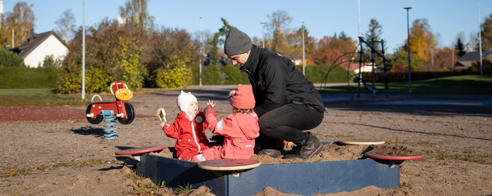 Nuori isä leikkii lastensa kanssa hiekkalaatikolla.