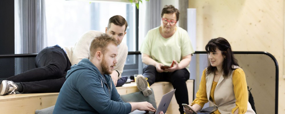 Työkavereiden porukka istuu toimistotilassa ja keskustelee kannettavan tietokoneen äärellä.