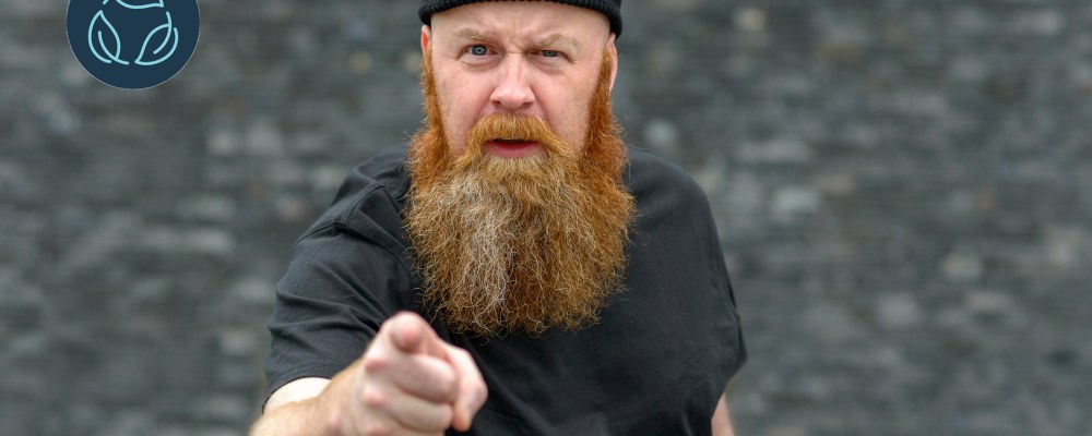A bearded person points at the camera, gray background.