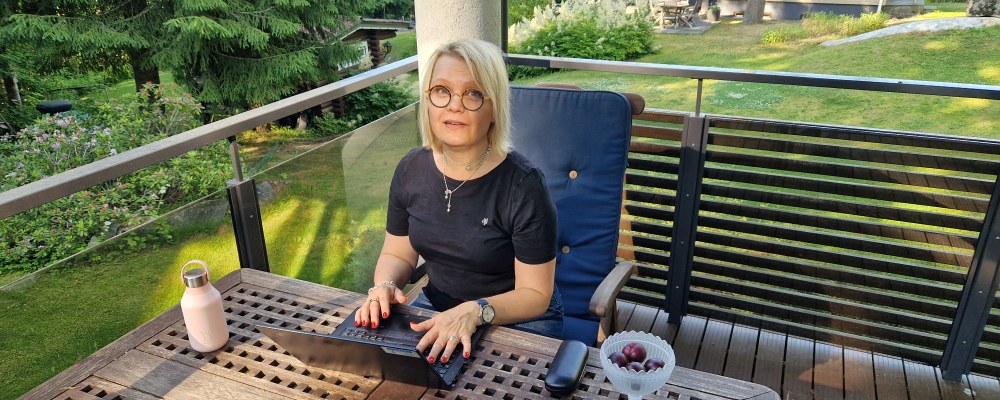Aino Mensonen istuu terassilla tietokoneen ääressä, takana puutarha.