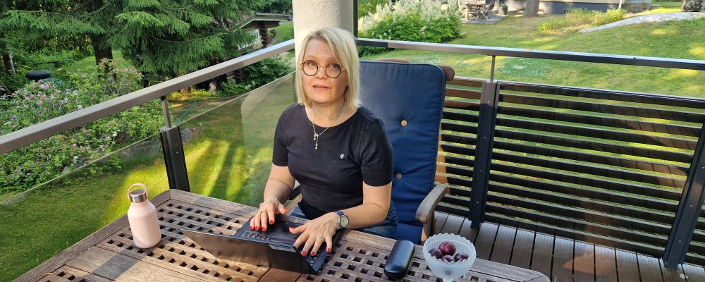 Aino Mensonen istuu terassilla tietokoneen ääressä, takana puutarha.