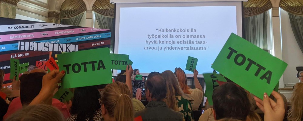 Pride-seminaarin yleisö on nostanut ilmaan vihreitä totta-lappuja, taustalla näkyy teksti: kaikenkokoisilla työpaikoilla on olemassa hyviä keinoja edistää tasa-arvoa ja yhdenvertaisuutta.