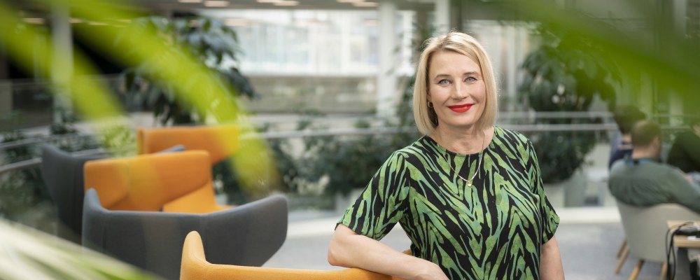 Pia Kautonen seisoo aulatilassa ja nojaa oranssiin tuoliin, etualalla on viherkasveja.