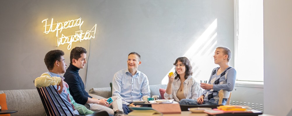 Viisi henkilöä juttelemassa pöydän ääressä, seinällä valotaulu jossa lukee "upeaa työtä"