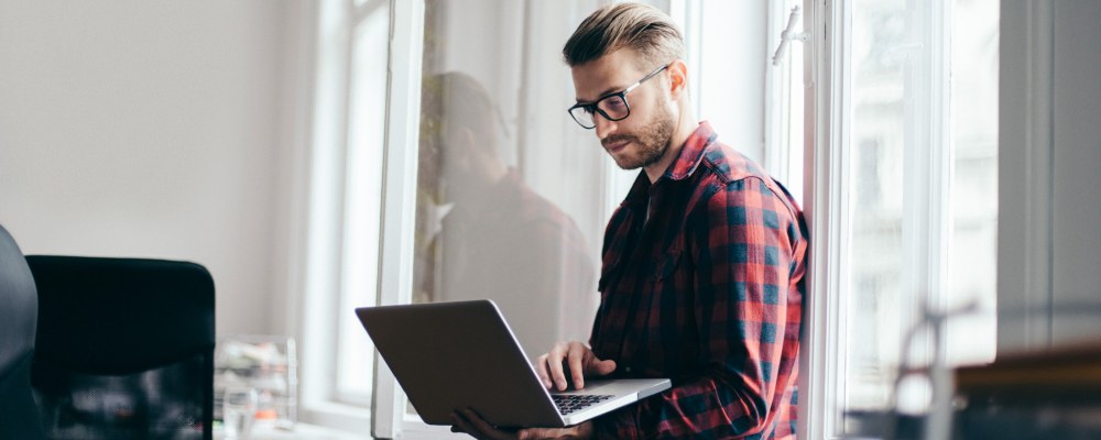 Ruutupaitainen mies istuu läppärin kanssa toimiston ikkunalaudalla.