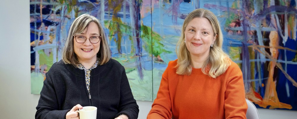 Satu Myller ja Maisa Holm istuvat pöydän ääressä, takana värikäs maalaus.
