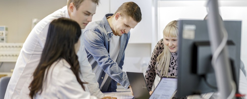 Ihmisiä kokoontuneena tietokoneiden ja papereiden ääreen työtilassa, osalla labratakit.