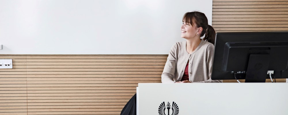 Reetta Kyynäräinen istuu luentosalissa tietokoneen ääressä ja katsoo sivulle. 