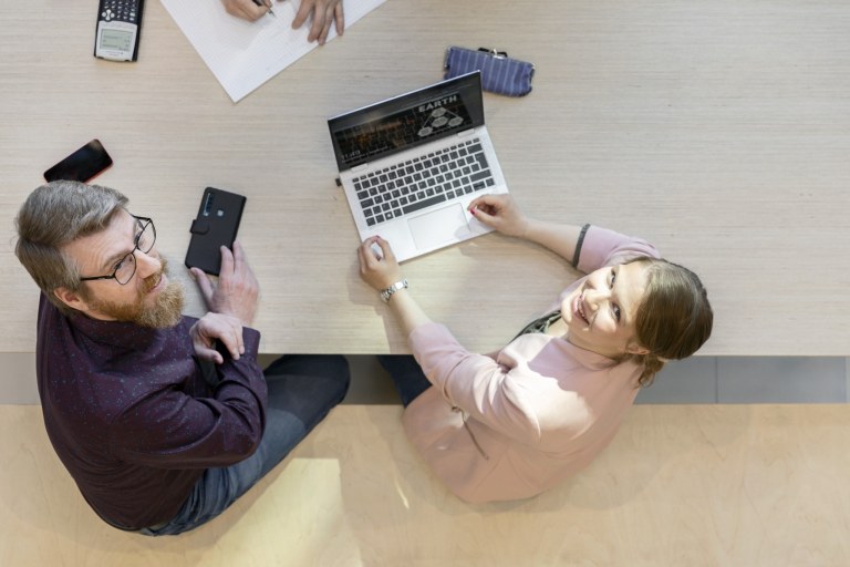 Kaksi henkilöä istuu tietokoneen ääressä ja katsoo kameraan.