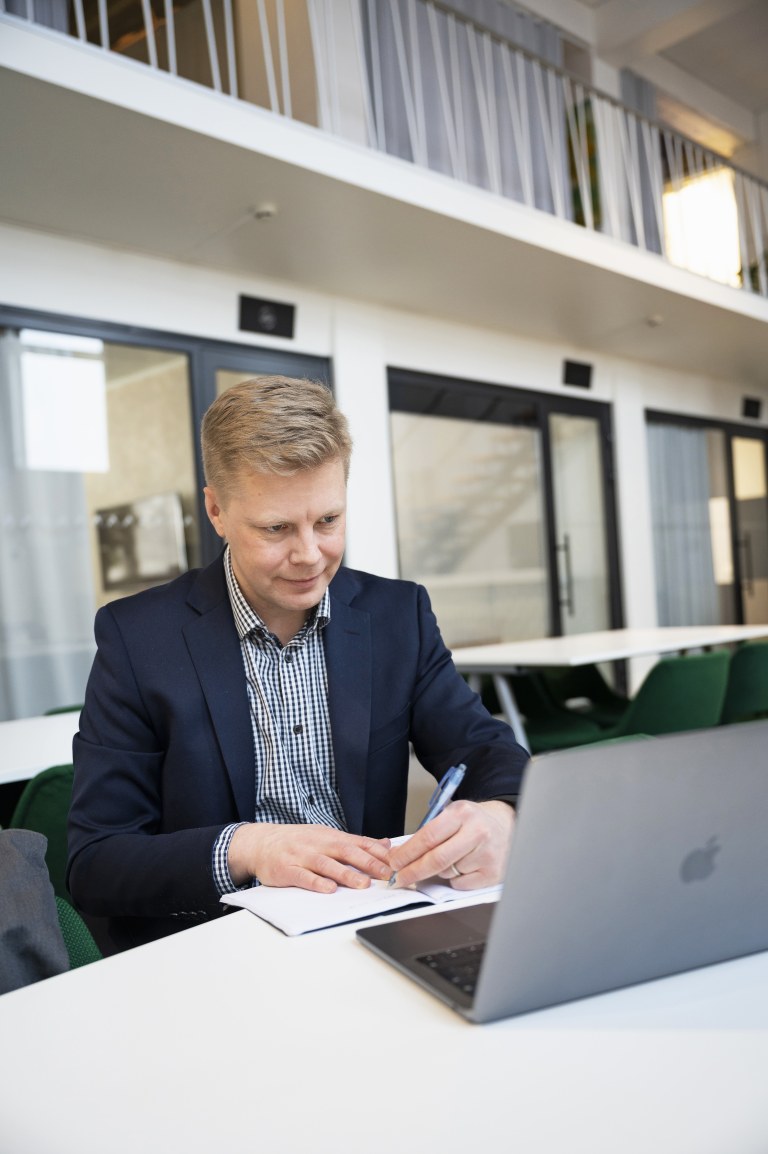 Tuomas Olkku lukee kannettavan tietokoneen näytöltä jotakin ja kirjoittaa samalla vihkoon.