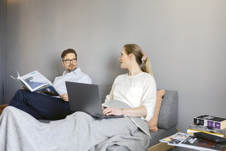 Kaksi henkilöä istuu sohvalla, toinen lukee lehteä ja toisella on tietokone edessään.