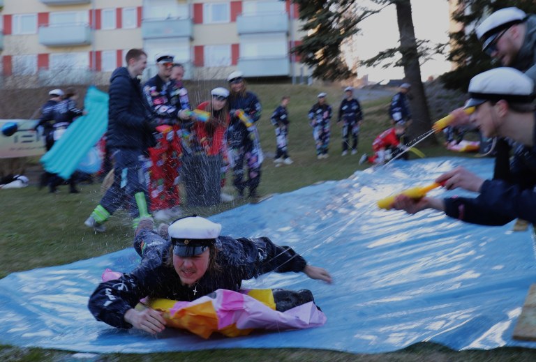 Teekkari laskee maata vasten laitetusta eristemuovista tehtyä liukumäkeä pitkin samalla kun muut ampuvat häntä kohden vesipyssyillä.