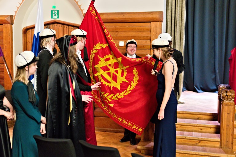 Juhla-asuisia ihmisiä teekkarilakit päässään lipun ympärillä.