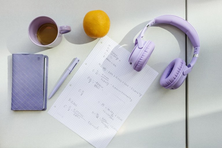 Pöydällä kuulokkeet, kahvikuppi, kalenteri, kynä, appelsiini ja paperi.