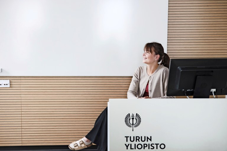 Reetta Kyynäräinen istuu luentosalissa tietokoneen ääressä ja katsoo sivulle. 
