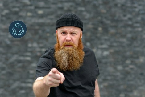 A bearded person points at the camera, gray background.