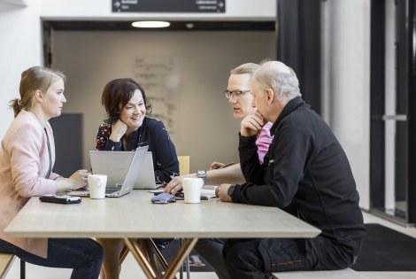 Neljä henkilöä istuu pöydän ääressä läppärin ja kahvikuppien kanssa.