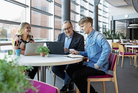 Kolme ihmistä istuu saman pöydän ääressä ruokalassa, edessään kaksi kannettavaa tietokonetta ja vihko.