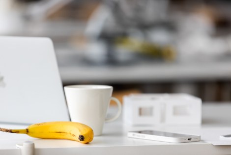 Pöydällä on tietokone, kahvikuppi, banaani ja älypuhelin