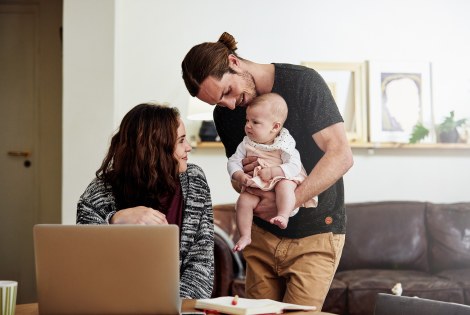 Vanhemmat vauvan kanssa, toinen vanhempi on tietokoneen ääressä ja toinen pitää vauvaa.