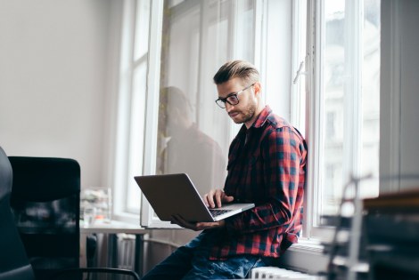 Ruutupaitainen mies istuu läppärin kanssa toimiston ikkunalaudalla.