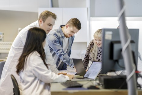 Ihmisiä kokoontuneena tietokoneiden ja papereiden ääreen työtilassa, osalla labratakit.