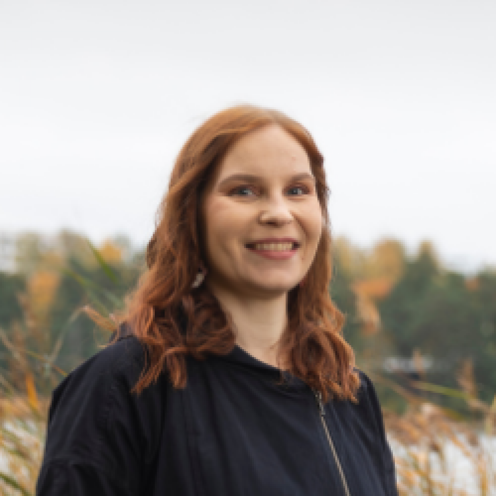 Kuvassa on ruskeahiuksinen nainen (Satu Ramsland), joka hymyilee. Taustalla näkyy lahti ja sen toisella puolella puita.