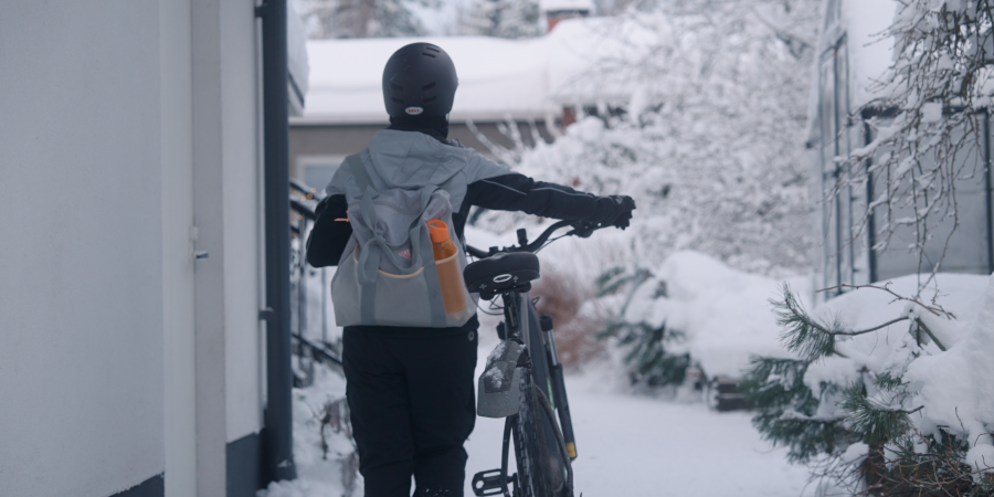 Ulkoiluasuun pukeutunut nainen taluttaa sähköavusteista pyörää kotipihalleen. Piha on luminen. 